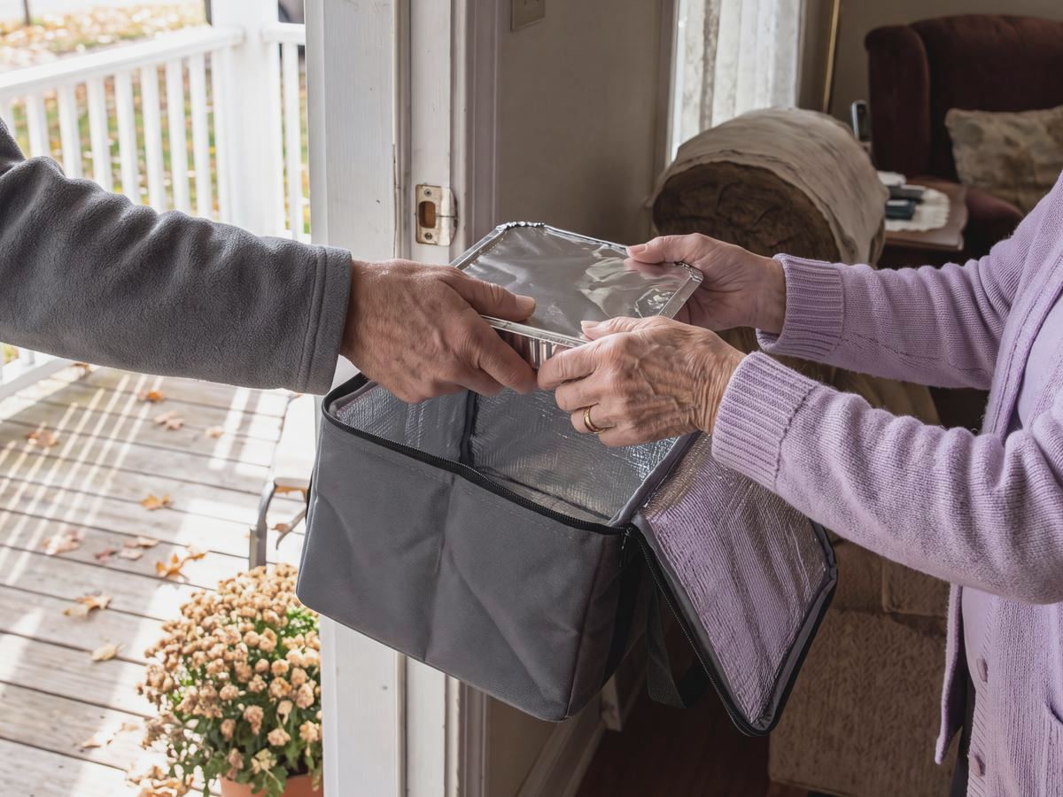 At an open front door, an older woman's hands take a covered meal tray from a volunteer's hands on an autumn morning.